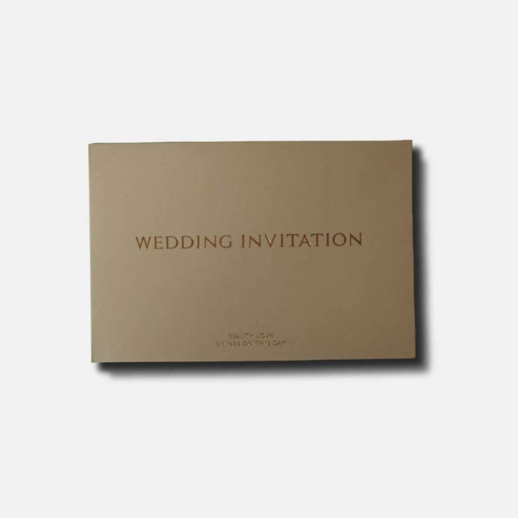 Elegant nude-colored wedding invitation envelope with gold embossed lettering stating 'WEDDING INVITATION' and a delicate script at the bottom reading 'Beauty, Love, Shines on This Day'.